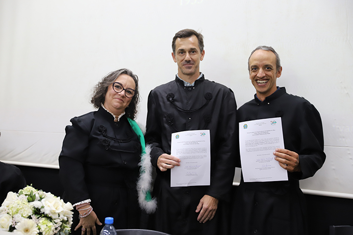 A imagem mostra três pessoas em trajes acadêmicos formais, em um ambiente de cerimônia oficial, possivelmente de posse. As duas pessoas à direita seguram documentos oficiais, enquanto a pessoa à esquerda (com faixa verde e detalhes em branco) posa ao lado deles com expressão solene. À frente, há um arranjo de flores brancas sobre a mesa. 