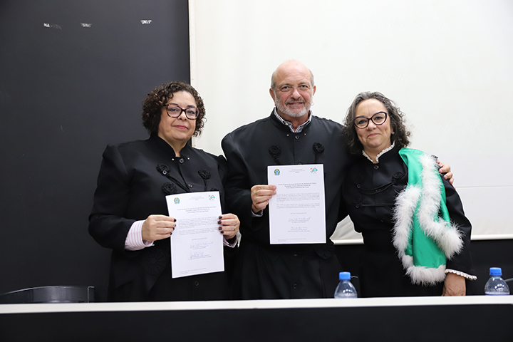 A imagem mostra três pessoas em trajes cerimoniais acadêmicos, segurando documentos oficiais. Elas estão sorrindo e posando para a foto em frente a uma mesa, sugerindo a formalização de uma posse ou nomeação. À direita, uma das pessoas veste uma faixa verde com detalhes em branco, tradicional em eventos de investidura. O ambiente é o mesmo auditório das imagens anteriores, da Universidade Federal de São Paulo (UNIFESP)