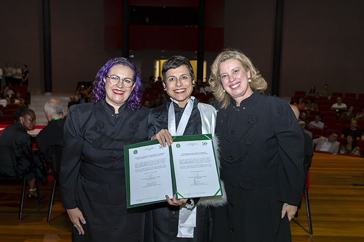 A imagem mostra três mulheres em um evento formal, possivelmente uma cerimônia de posse ou formatura. Elas estão vestindo trajes pretos formais, sendo que a pessoa no centro também usa uma faixa branca sobre a roupa e segura um certificado ou diploma com moldura verde. Todas estão sorrindo e parecem felizes com a ocasião. O fundo da imagem revela um auditório com pessoas sentadas e outras de pé, indicando que se trata de um evento oficial. A iluminação e o ambiente sugerem um espaço acadêmico ou institucional.