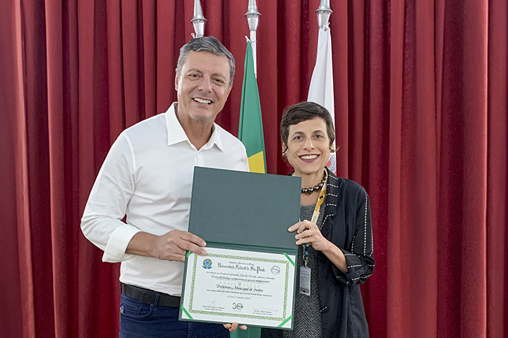 A imagem mostra um homem e uma mulher sorrindo enquanto seguram um certificado. O homem, à esquerda, veste uma camisa branca de mangas compridas e calça jeans escura. A mulher, à direita, veste um blazer preto sobre uma blusa escura e usa um crachá pendurado no pescoço. Atrás deles, há uma cortina vermelha e duas bandeiras hasteadas, uma das quais parece ser a bandeira do Brasil. O certificado que seguram tem uma moldura verde e inclui um logotipo oficial. Eles parecem estar participando de uma cerimônia de premiação ou reconhecimento em um ambiente formal.