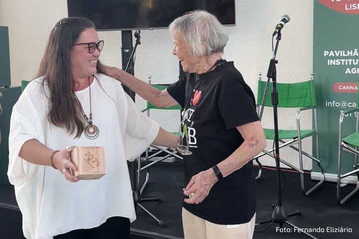 A imagem mostra duas mulheres sorrindo e se abraçando em um palco. A mulher à esquerda segura um troféu de madeira com a inscrição "2023" e usa uma blusa branca. A mulher à direita, de cabelo grisalho curto, veste uma camiseta preta com a frase "I ❤️ CBD". Ao fundo, há microfones, cadeiras verdes e um banner com informações do evento. A foto é creditada a Fernando Elizário.