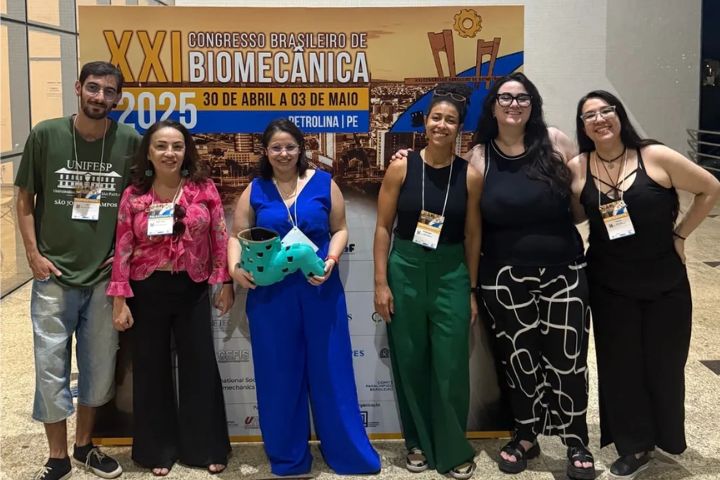A imagem mostra um grupo de seis pessoas posando em frente a um banner de evento. O banner anuncia o "XXI Congresso Brasileiro de Biomecânica", que acontecerá de 30 de abril a 03 de maio de 2025, em Petrolina - PE. O fundo do banner inclui uma imagem da cidade e elementos gráficos que remetem à biomecânica e à engenharia.  As pessoas estão sorrindo e aparentam estar participando do congresso. Elas usam crachás de identificação, indicando que são participantes ou palestrantes do evento. No centro do grupo, uma mulher de macacão azul segura uma órtese de membro inferior, o que pode indicar que o grupo está relacionado a algum projeto ou apresentação sobre órteses.