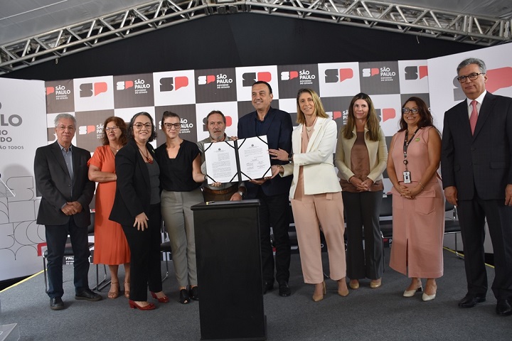 A imagem mostra um grupo de dez pessoas reunidas em um evento oficial, realizado sob uma estrutura coberta. No centro, duas pessoas seguram um documento aberto, simbolizando a assinatura de um protocolo de intenções.  O fundo contém um painel com a logomarca do estado de São Paulo e a frase "Governo de São Paulo - Um Estado de Todos", indicando que o evento está relacionado a ações governamentais.  Os participantes vestem trajes formais e estão organizados em uma pose solene, demonstrando a importância do momento. O evento parece envolver autoridades e representantes de instituições em uma cerimônia de assinatura de um compromisso ou parceria.