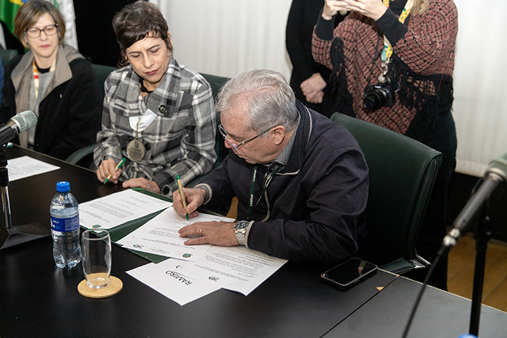 A imagem mostra um momento de assinatura formal de documentos. Um homem de cabelos grisalhos, sentado à mesa, está assinando os papéis com uma caneta verde, enquanto uma mulher ao seu lado observa atentamente. Ambos usam crachás, indicando participação em um evento oficial. Outras pessoas estão ao fundo, uma delas segurando uma câmera fotográfica, registrando o momento. A mesa está equipada com microfones, copos e garrafa de água, sugerindo um ambiente de conferência ou cerimônia institucional.