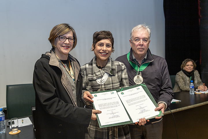 A imagem mostra três pessoas em pé, sorrindo e segurando juntas dois documentos abertos, aparentemente certificados ou termos de compromisso. Eles estão em um ambiente formal, provavelmente uma cerimônia ou evento institucional. Ao fundo, uma mulher sentada observa a cena. Todos estão bem vestidos e usam crachás, indicando participação oficial no evento.