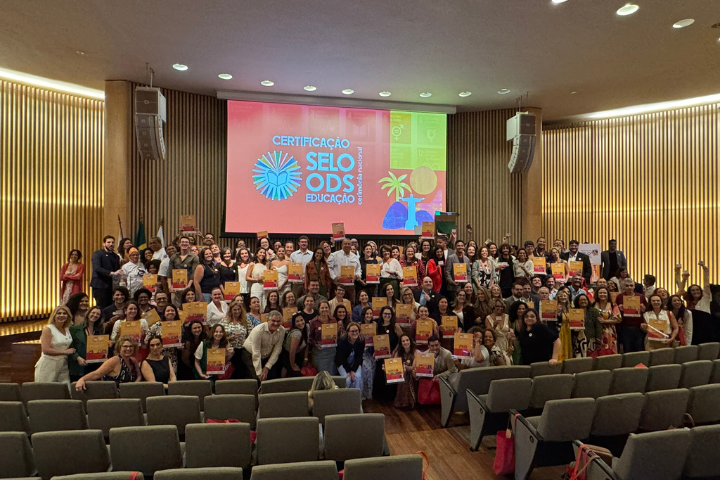 A imagem mostra um grande grupo de pessoas reunidas em um auditório para um evento de certificação do Selo ODS Educação. No fundo, um telão exibe um design colorido com o texto "CERTIFICAÇÃO SELO ODS EDUCAÇÃO", junto a elementos gráficos como um sol estilizado e ícones dos Objetivos de Desenvolvimento Sustentável (ODS). Os participantes estão sorrindo e segurando certificados semelhantes, sugerindo que receberam um reconhecimento ou premiação. O auditório tem um design moderno, com cadeiras acolchoadas, paredes de madeira e iluminação agradável. A diversidade de pessoas na foto, incluindo diferentes idades e estilos de vestimenta, indica um evento inclusivo e colaborativo. Muitas delas parecem representar instituições educacionais ou projetos alinhados com os ODS. A atmosfera geral transmite um sentimento de celebração e conquista.