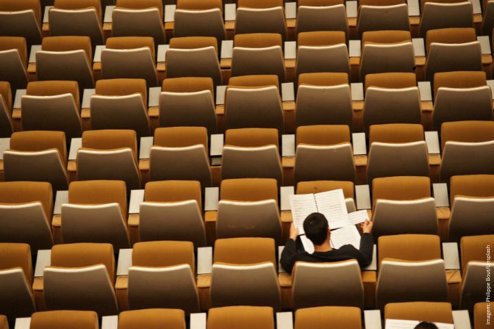 A imagem mostra um auditório com muitas cadeiras vazias, organizadas em fileiras simétricas. No meio delas, há uma única pessoa sentada, vestindo uma roupa escura, segurando e lendo uma partitura musical. A composição da imagem enfatiza a solidão do indivíduo em meio ao grande espaço vazio, criando uma sensação de isolamento ou concentração profunda. A paleta de cores é neutra, com tons de marrom e bege predominando.