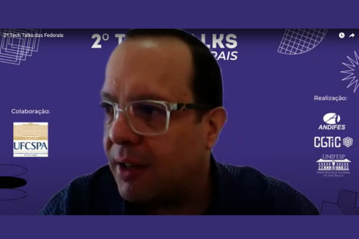 A imagem mostra um homem de óculos participando de uma videoconferência, com um fundo virtual roxo que exibe logotipos de organizações como UFCSPA (colaboração), ANDIFES, CGTIC e UNIFESP (realização), além do título do evento “2º Tech Talks das Federais”.