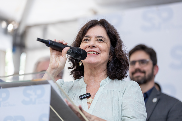 A imagem mostra uma mulher de cabelos escuros e ondulados falando ao microfone com um sorriso no rosto. Ela está vestindo uma blusa clara e usa um pequeno microfone de lapela preso à roupa. Atrás dela, há um fundo com o logotipo "SP" repetido, sugerindo um evento oficial ou institucional. Algumas pessoas estão desfocadas ao fundo, incluindo um homem de terno e óculos, que parece estar acompanhando a fala dela. O clima da imagem transmite positividade e entusiasmo, indicando um momento importante e celebrado.