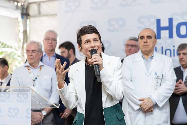 A imagem mostra uma mulher de cabelos curtos discursando em um evento. Ela segura um microfone e faz um gesto com a mão, parecendo expressiva e engajada. Ela veste um blazer branco com detalhes verdes e uma blusa preta.  Atrás dela, há várias pessoas, incluindo médicos de jaleco branco e outras figuras de terno, que aparentam ser profissionais da área da saúde e autoridades. O fundo exibe um banner com o logotipo "SP" e as palavras "Hospital" e "Unidade de Emergência Referenciada", indicando que o evento está relacionado à saúde pública, possivelmente uma inauguração ou anúncio importante.  O ambiente tem um clima formal e institucional, e a expressão da mulher sugere entusiasmo e comprometimento com o que está sendo apresentado.