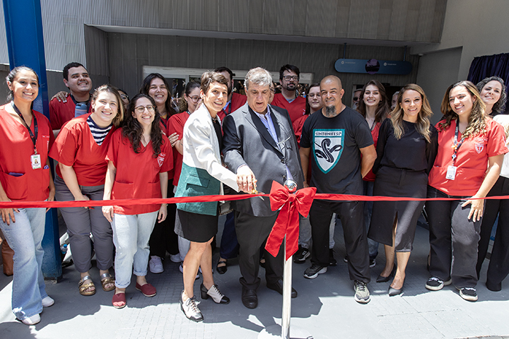 A imagem mostra um grupo de pessoas reunidas para uma cerimônia de inauguração. No centro, um homem de terno e uma mulher de blazer branco e saia preta seguram juntos uma tesoura, cortando uma fita vermelha com um laço decorativo, simbolizando a abertura oficial de um novo espaço. Ao redor deles, há várias pessoas sorrindo e posando para a foto, muitas vestindo jalecos vermelhos, possivelmente indicando que fazem parte de uma equipe de saúde ou acadêmica. Algumas pessoas usam crachás de identificação, e o ambiente sugere a entrada de um edifício institucional. O clima da foto é festivo e positivo.