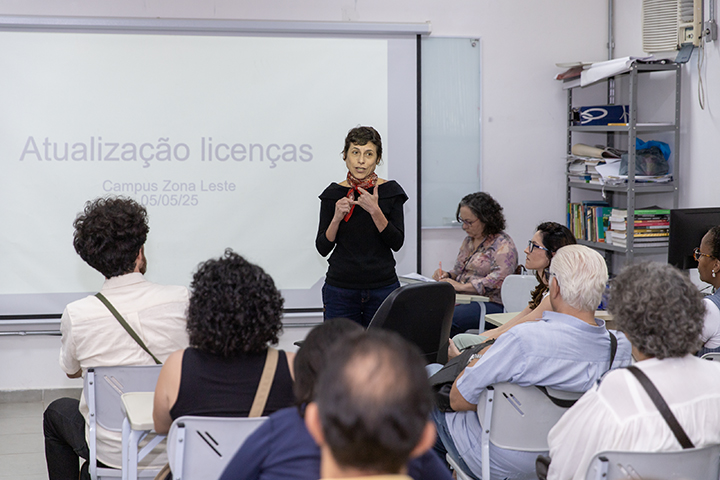 A imagem mostra uma sala de aula ou auditório pequeno, onde está ocorrendo uma apresentação ou palestra. No centro da imagem, uma mulher está em pé, falando para o público. Ela está vestida com roupas escuras e usa um lenço vermelho no pescoço. Atrás dela, há um projetor exibindo um slide com o título "Atualização licenças" e, logo abaixo, o texto "Campus Zona Leste 05/05/25".  O público está sentado em cadeiras, de frente para a apresentadora. A maioria das pessoas está prestando atenção, e algumas fazem anotações. No fundo, do lado direito da imagem, há prateleiras com livros e materiais diversos, além de um ar-condicionado fixado na parede. A atmosfera parece ser de um ambiente educacional ou institucional, com foco em capacitação ou atualização profissional.