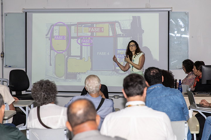 Esta imagem retrata uma apresentação técnica ou reunião institucional com foco em planejamento ou arquitetura de espaços, possivelmente vinculada à Unifesp.  Elementos da imagem: Apresentadora: Uma mulher está em pé à frente da sala, explicando um mapa esquemático projetado na tela, que exibe divisões por “FASE 1”, “FASE 3” e “FASE 4”, indicando prováveis etapas de construção ou reforma.  Público: Vários participantes, sentados de frente para a tela, acompanham atentamente.  Ambiente: Sala de aula ou reunião, equipada com projetor, quadro branco e cadeiras organizadas em fileiras.