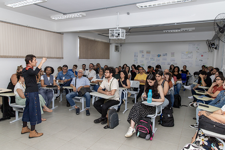 A imagem mostra uma sala de aula cheia, com diversas pessoas sentadas em carteiras escolares, todas voltadas para a frente, onde uma mulher está em pé conduzindo uma apresentação ou palestra. Ela é a mesma da imagem anterior: veste roupas escuras, usa um lenço vermelho no pescoço e botas marrons. A mulher está gesticulando com a mão direita, aparentemente enfatizando um ponto para o público.  Os participantes parecem ser adultos, indicando que o evento é voltado a um público profissional ou universitário. Muitos estão atentos, alguns fazem anotações ou observam a palestrante com interesse. Há mochilas e garrafas de água sobre algumas carteiras, e a parede ao fundo exibe vários cartazes e folhas com anotações ou diagramas, sugerindo que a sala é usada para fins educacionais ou acadêmicos.  O ambiente é bem iluminado, com luzes fluorescentes no teto, e há ventiladores e ar-condicionado, indicando uma preocupação com o conforto dos presentes. A atmosfera geral é de um evento educacional, como um curso de formação, capacitação ou uma reunião pedagógica.