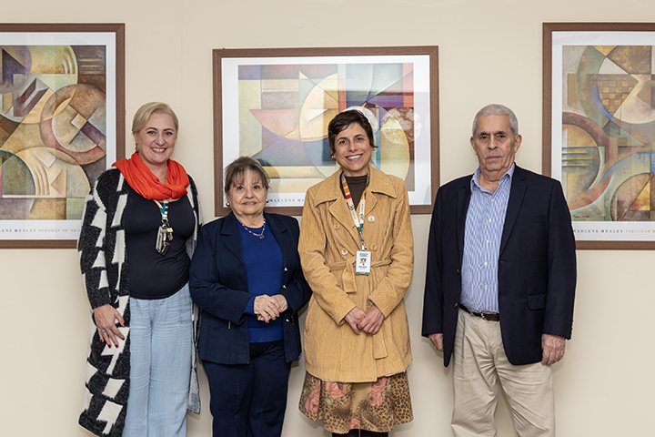A imagem mostra quatro pessoas em pé, sorrindo, diante de uma parede com três quadros de arte abstrata. São três mulheres e um homem, todos vestidos de forma formal ou semiformal, sugerindo um ambiente institucional ou profissional.