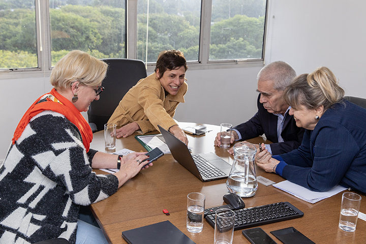 A imagem mostra as mesmas quatro pessoas da foto anterior reunidas em uma sala de reuniões. Elas estão sentadas ao redor de uma mesa de madeira, interagindo com um notebook. A mulher de casaco bege aponta para a tela, enquanto os outros observam com atenção e sorrisos, sugerindo um clima colaborativo e descontraído. Há copos de água, uma jarra, celulares e cadernos sobre a mesa. Ao fundo, janelas amplas mostram uma vista arborizada.