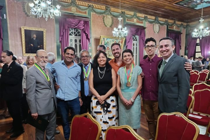 A imagem mostra um grupo de oito pessoas reunidas em um salão elegante com decoração clássica. O ambiente possui cortinas roxas, lustres antigos e retratos nas paredes, sugerindo um espaço histórico ou institucional. As cadeiras vermelhas e douradas indicam que o local foi preparado para um evento formal.  Entre os presentes, duas pessoas estão usando medalhas com fitas verde, amarela e vermelha, o que sugere que foram homenageadas na cerimônia. Uma dessas pessoas é uma mulher vestindo um vestido verde claro, sorrindo ao lado dos demais. O grupo é composto por homens e mulheres de diferentes idades, todos sorridentes e posando para a foto, demonstrando um clima de confraternização e celebração.  Ao fundo, outras pessoas podem ser vistas conversando e circulando pelo salão, reforçando a ideia de um evento social ou de premiação.