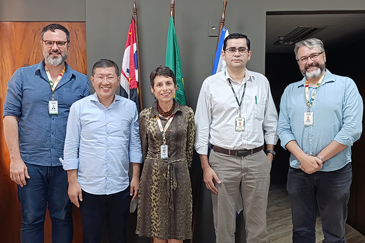 A imagem mostra cinco pessoas posando para uma foto em um ambiente institucional. Ao fundo, há três bandeiras, incluindo a do Brasil e a do estado de São Paulo, fixadas em suportes de madeira. Os indivíduos estão vestindo roupas sociais ou semi-formais, e todos usam crachás, sugerindo que fazem parte de uma equipe de trabalho ou evento oficial. Eles estão sorrindo e aparentam estar confortáveis, indicando um momento de registro de reunião ou evento profissional. O fundo conta com uma parede de cor escura, uma porta de madeira e um corredor levemente iluminado.