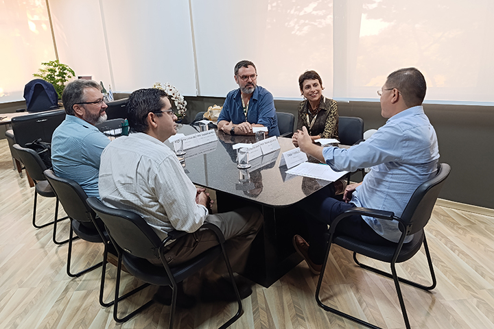 A imagem mostra cinco pessoas reunidas ao redor de uma mesa em um ambiente corporativo ou institucional. A mesa tem uma superfície preta brilhante e está organizada com plaquinhas de identificação, copos de água e papéis, sugerindo uma reunião formal.  Os participantes estão vestidos com trajes sociais ou semi-formais e usam crachás, o que indica que fazem parte de uma equipe profissional. A conversa parece envolvente, com um dos homens gesticulando enquanto fala, enquanto os outros ouvem atentamente.  O ambiente é bem iluminado, com grandes janelas cobertas por cortinas translúcidas, permitindo a entrada de luz natural. O chão tem um acabamento de madeira clara, e o fundo conta com um sofá e uma mesa decorada com plantas e objetos.