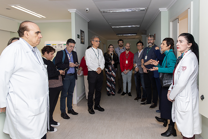 Nesta imagem, o grupo de visitantes está novamente em um corredor de ambiente hospitalar, ouvindo explicações de dois profissionais de saúde — uma mulher e um homem, ambos usando jaleco branco. A mulher de jaleco parece estar detalhando informações, enquanto o grupo observa atentamente.  Os visitantes, identificáveis pelos crachás, estão dispostos em semicírculo, o que sugere uma apresentação ou uma visita guiada estruturada. Vemos algumas pessoas fazendo anotações, indicando que a visita tem caráter técnico ou de avaliação. A mulher de blazer verde-água, que apareceu em imagens anteriores, também participa ativamente da conversa.