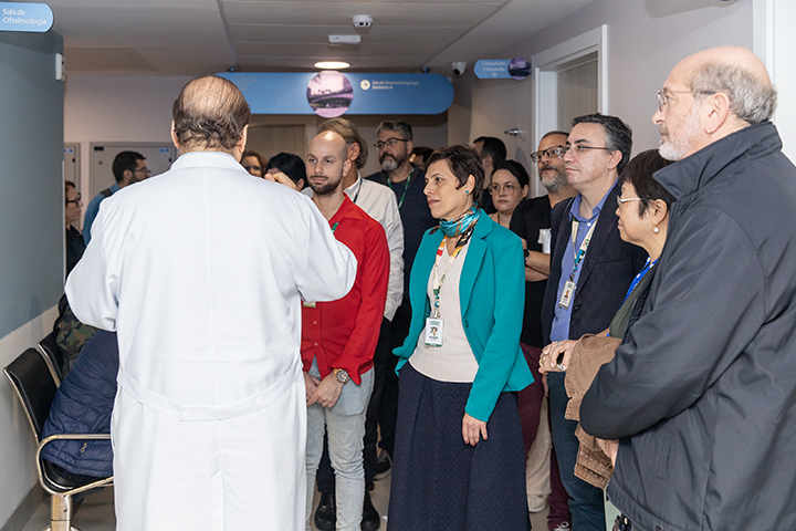 Nesta imagem, vemos um grupo de pessoas reunido em um corredor de hospital ou instituição de saúde, acompanhando atentamente a explicação de um profissional da saúde — um homem de jaleco branco que está de costas para a câmera, aparentemente conduzindo uma visita guiada ou explicação técnica.  Os visitantes, muitos com crachás, estão atentos ao que é falado. Alguns dos rostos são os mesmos que aparecem nas imagens anteriores, o que sugere que se trata de uma continuidade daquele evento ou visita institucional. À esquerda da imagem, há uma placa indicando a "Sala de Oftalmologia", reforçando que o grupo está em um setor especializado.