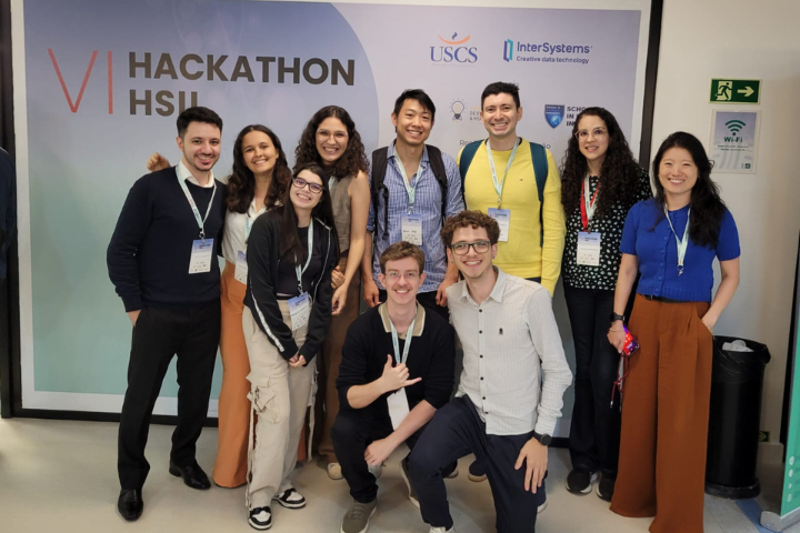 A imagem mostra um grupo de jovens participantes posando em frente a um painel do evento “VI Hackathon HSI”, realizado pela USCS em parceria com a InterSystems e outras instituições. Todos usam crachás de identificação, indicando que fazem parte oficial do evento. O grupo aparenta estar animado e unido, representando um momento de confraternização e celebração em um ambiente de inovação e tecnologia voltado à saúde.