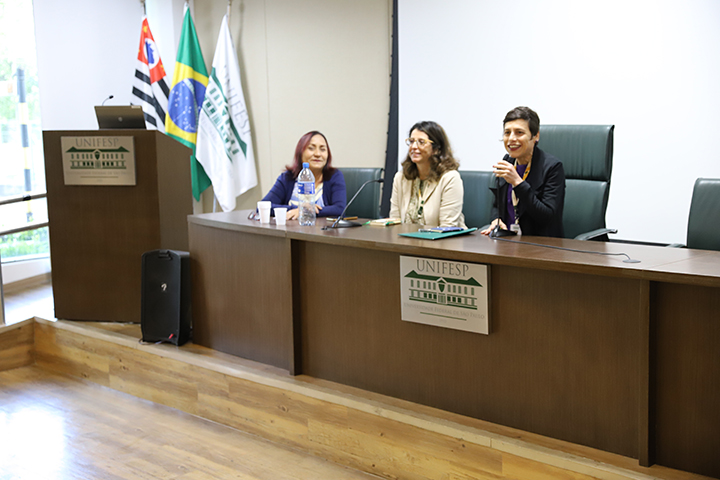 A imagem mostra três mulheres sentadas atrás de uma mesa em um auditório da UNIFESP (Universidade Federal de São Paulo), participando de um evento ou reunião. Ao fundo, há bandeiras do Brasil, do estado de São Paulo e da própria universidade. As participantes parecem engajadas e sorridentes, sugerindo um ambiente formal, porém acolhedor.