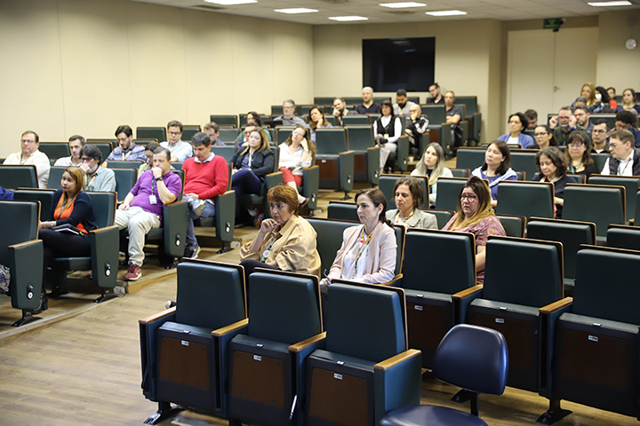A imagem mostra um auditório parcialmente cheio, com dezenas de pessoas sentadas em cadeiras acolchoadas, assistindo atentamente a um evento. O ambiente é formal, com público adulto, sugerindo uma conferência, palestra ou reunião acadêmica. A maioria das pessoas aparenta estar focada na apresentação à frente, fora do campo de visão da imagem.