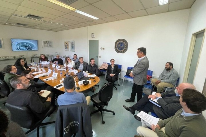 Representantes das seis universidades públicas paulistas sentados em uma mesa assistindo a uma apresentação feita por um homem de paletó cinza e calça preta