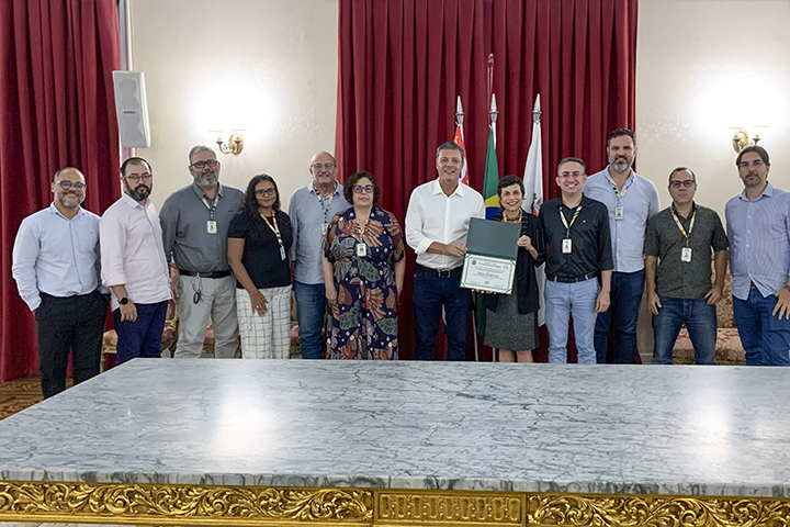 A imagem mostra um grupo de 12 pessoas posando para uma foto em um salão com cortinas vermelhas e bandeiras ao fundo. No centro, um homem e uma mulher seguram um certificado dentro de uma pasta verde, sugerindo uma cerimônia de premiação ou reconhecimento. A mulher segura o certificado com um sorriso. Os participantes estão vestidos com trajes sociais ou semi-formais, e alguns usam crachás. A mesa em primeiro plano tem um tampo de mármore e detalhes dourados na borda, indicando um ambiente institucional ou governamental. A iluminação vem de lustres nas paredes.