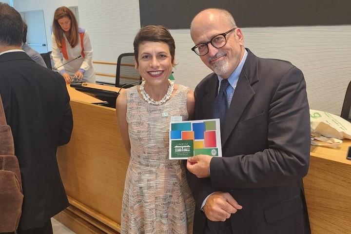 Nesta imagem, vemos uma mulher e um homem posando juntos, sorridentes, em um ambiente interno e moderno, provavelmente uma sala de eventos ou recepção. A mulher veste um vestido claro estampado e um colar branco, enquanto o homem está de terno e gravata, usando óculos. O cenário ao fundo inclui uma bancada de madeira e outras pessoas envolvidas em atividades, sugerindo que este momento aconteceu durante um evento ou conferência. A atmosfera é amistosa e descontraída, mas ainda formal, compatível com eventos institucionais ou diplomáticos.