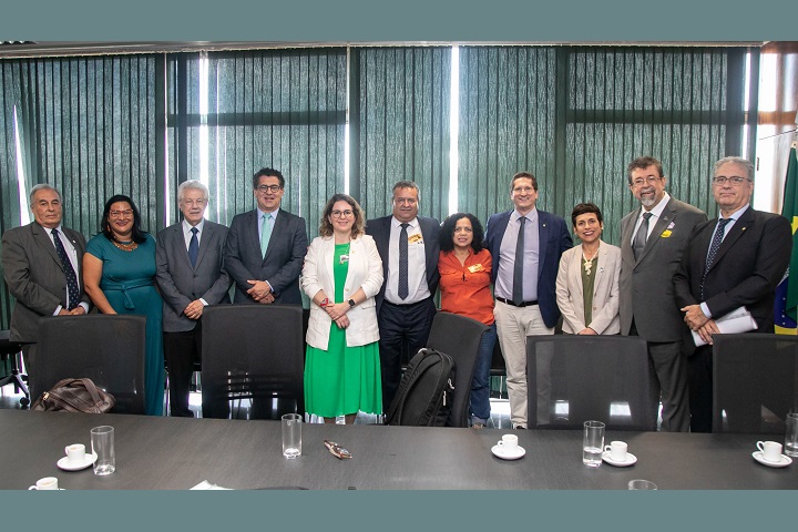 A imagem mostra um grupo de pessoas posando para uma foto em uma sala de reuniões. Ao todo, há onze pessoas, a maioria vestida formalmente, com roupas sociais. Algumas mulheres no grupo estão usando vestidos ou blazers. O fundo apresenta cortinas verticais de cor escura, que cobrem janelas grandes, deixando entrar um pouco de luz natural. Sobre a mesa, em primeiro plano, estão algumas xícaras de café e copos d'água. A disposição sugere um encontro oficial ou uma reunião de trabalho.