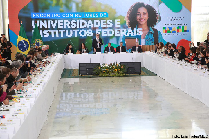A imagem mostra uma ampla reunião oficial do Ministério da Educação (MEC) do Brasil, com o título destacado ao fundo: "Encontro com Reitores de Universidades e Institutos Federais". O evento é promovido pelo Governo Federal, conforme indicado pelos logotipos no painel de fundo, que também exibe a identidade visual do governo com os dizeres “União e Reconstrução”.  O local é bem iluminado, com piso de mármore e uma grande mesa em formato de "U", onde dezenas de pessoas estão sentadas, incluindo reitores, autoridades do MEC e representantes institucionais. Ao centro, na cabeceira da mesa, está o ministro da Educação (provavelmente em pé, discursando), ladeado por outros membros do governo. Atrás dele estão as bandeiras do Brasil e da República, reforçando o caráter oficial da ocasião.  Na parte inferior da imagem, novamente se vê um arranjo floral com girassóis. As placas de identificação e o ambiente solene sugerem que o tema do encontro é relevante — como orçamento, políticas públicas, financiamento ou planejamento estratégico das instituições federais de ensino.  O crédito da imagem é de Luís Fortes/MEC, o que confirma o registro institucional do evento.