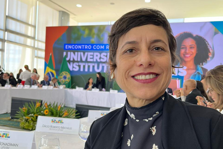 A imagem mostra uma mulher sorridente em primeiro plano, aparentemente tirando uma selfie durante um evento oficial. Ela tem cabelos curtos castanhos escuros e está vestindo uma blusa preta com detalhes brancos e um blazer preto.  Ao fundo, há uma mesa longa com várias pessoas sentadas, sugerindo uma reunião ou conferência formal. Atrás da mesa, há um grande painel colorido com os dizeres “ENCONTRO COM REITORES UNIVERSIDADES E INSTITUTOS”, indicando que o evento está relacionado ao Ministério da Educação (MEC) do Brasil e envolve representantes de instituições de ensino superior. Também é possível ver bandeiras do Brasil e o brasão da República, reforçando o caráter institucional da reunião.