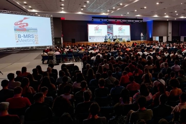 A imagem mostra um grande auditório repleto de pessoas assistindo a uma apresentação em um palco. O auditório está lotado, com o público sentado em fileiras de cadeiras, voltados para o palco onde há um painel de discussão em andamento. Duas grandes telas de projeção exibem o logotipo do evento e informações relacionadas.  Na tela à esquerda, lê-se: XXIII B-MRS Meeting 2024, sugerindo que o evento é o encontro anual da Sociedade Brasileira de Pesquisa em Materiais (B-MRS). O local parece ser um centro de convenções, com uma estrutura ampla e profissional, incluindo iluminação de palco e um grande espaço para o público.