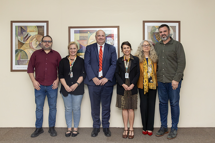 A imagem mostra um grupo de seis pessoas posando para uma foto em ambiente interno, diante de uma parede com três quadros de arte abstrata. O grupo é composto por três mulheres e três homens, todos de pé, lado a lado, sorrindo para a câmera em postura formal e amigável. Algumas das pessoas usam crachás institucionais, indicando participação em um encontro oficial ou reunião de trabalho. Ao centro está um homem usando terno azul escuro, camisa branca e gravata vermelha. À sua esquerda e direita estão as demais pessoas, vestidas de forma variada, entre roupas sociais e casuais. O contexto, a composição da foto e os trajes sugerem que se trata de uma visita institucional ou diplomática. Se quiser, posso te ajudar a escrever um texto para publicação institucional, criar uma legenda inclusiva com descrição de acessibilidade, ou compor uma matéria sobre essa reunião ou visita.