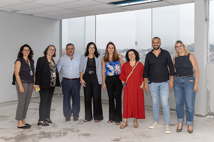 A imagem mostra um grupo de oito pessoas posando para uma foto em um ambiente interno, aparentemente um espaço corporativo ou acadêmico em fase de finalização, devido ao chão e paredes ainda inacabados. O grupo é composto por homens e mulheres vestidos de maneira profissional e casual, sugerindo um evento institucional. O fundo conta com janelas de vidro, permitindo uma vista da cidade ao fundo. Todos parecem estar sorrindo e olhando para a câmera, indicando um registro formal ou comemorativo.