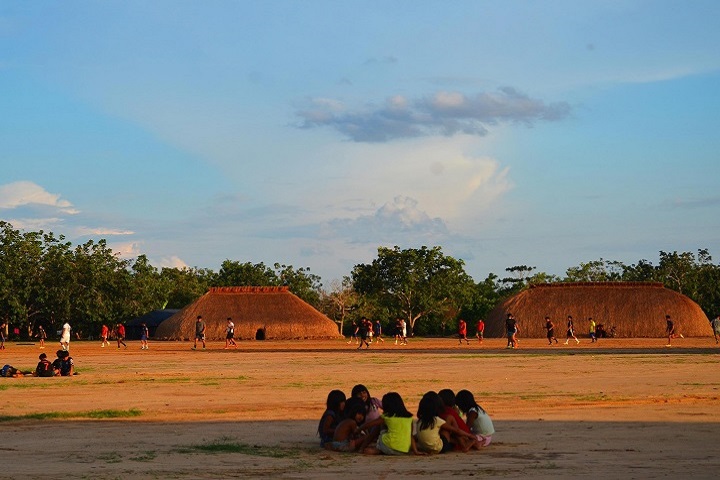 A imagem captura uma cena vibrante e culturalmente rica em uma aldeia indígena. No centro, um amplo espaço aberto de terra batida serve como palco para diversas atividades. Crianças indígenas, vestindo roupas coloridas e tradicionais, estão reunidas em um círculo, observando atentamente algo que ocorre ao fundo. Seus rostos transmitem curiosidade e animação.  Ao fundo, a paisagem se estende com casas típicas das comunidades indígenas, construídas com materiais naturais e apresentando formas cônicas características. Essas casas, distribuídas em um campo aberto, criam um cenário bucólico e convidativo. A vegetação circundante, composta por árvores altas e arbustos, adiciona um toque de verde à composição e proporciona sombra aos moradores da aldeia.  O céu, um azul claro e límpido, contrasta com as nuvens brancas e esponjosas que se espalham pelo horizonte. A luz natural do sol, que incide sobre a cena, confere à imagem um tom quente e acolhedor, realçando as cores vibrantes das roupas das crianças e da vegetação.