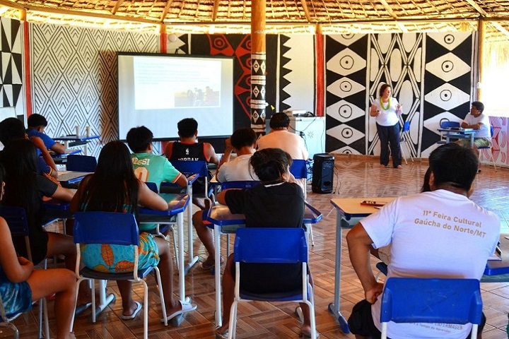 A imagem mostra um grupo de pessoas participando de uma aula ou oficina em um ambiente tradicional, semelhante a uma oca, com decoração típica indígena nas paredes, como padrões geométricos em preto, branco e vermelho. Os participantes estão sentados em carteiras escolares, prestando atenção a uma apresentação. Uma mulher está de pé, aparentemente conduzindo a atividade, com um projetor exibindo uma imagem em uma tela à frente. A atmosfera sugere um evento educacional, como uma oficina ou curso voltado para a comunidade local, combinando saberes tradicionais e conhecimentos externos.