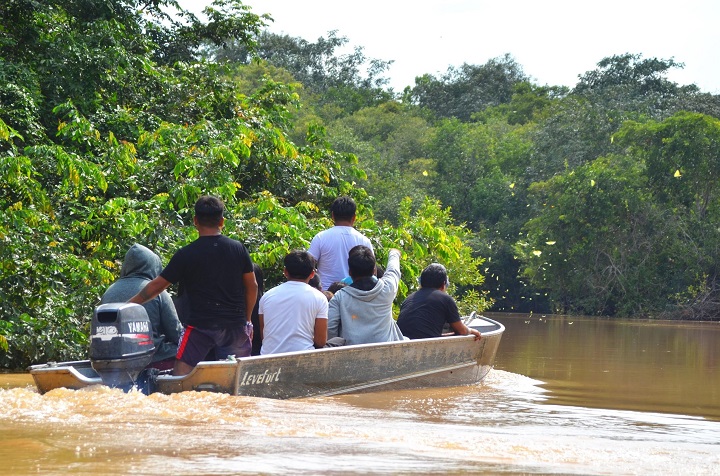 A imagem mostra um grupo de pessoas em um barco motorizado navegando em um rio de águas turvas, cercado por vegetação densa e verde, sugerindo uma floresta tropical. O motor do barco é visível na parte traseira, com a marca "Yamaha" destacada. O grupo está sentado, com alguns membros apontando para algo no ambiente ao redor, possivelmente observando a fauna ou a paisagem. A cena sugere um passeio ou expedição em um ambiente natural, como a Amazônia ou outro rio em área de floresta tropical.