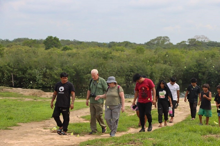 A imagem mostra um grupo de pessoas caminhando por uma trilha de terra em um ambiente rural ou natural, cercado por vegetação densa, provavelmente uma floresta tropical. O grupo inclui adultos e crianças, alguns vestindo roupas casuais e outros usando trajes que parecem típicos ou culturais. Entre eles, há pessoas carregando mochilas e objetos como garrafas de água. O clima parece ensolarado, e o terreno é coberto por grama em algumas áreas. A cena sugere uma caminhada ou atividade comunitária, possivelmente em uma área preservada ou indígena.