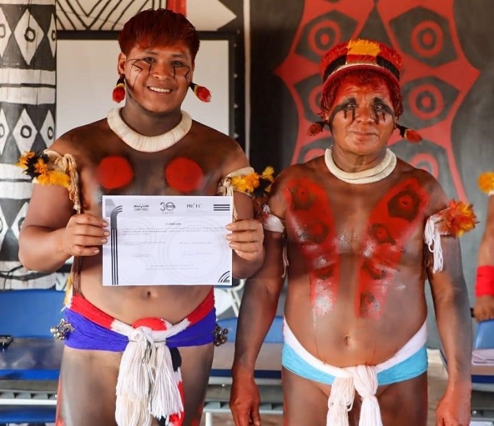 A imagem mostra dois homens vestidos com trajes tradicionais indígenas. Ambos estão com o corpo pintado com padrões simétricos em vermelho e preto, adornados com acessórios feitos de penas coloridas e colares. O homem à esquerda sorri e segura um certificado ou documento, enquanto o homem à direita está em pé ao lado dele, em uma postura séria. Ao fundo, há elementos decorativos com desenhos geométricos e cores predominantes em preto, vermelho e branco, possivelmente representando a cultura indígena. A cena parece estar relacionada a uma celebração ou cerimônia formal, talvez marcando a conclusão de um curso, projeto ou outro tipo de reconhecimento.