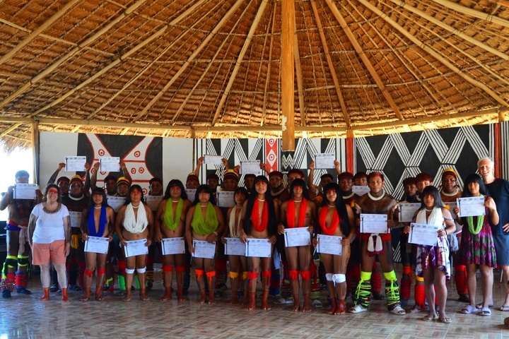 A imagem mostra um grupo de pessoas reunidas em um ambiente tradicional, provavelmente uma oca, com teto de palha e elementos decorativos típicos de uma cultura indígena. O grupo é composto por homens e mulheres, alguns vestindo trajes e adornos tradicionais coloridos, como colares, cintos, faixas e pintura corporal, que refletem a cultura do povo Wauja. Muitas pessoas seguram certificados, indicando que participaram de alguma atividade ou curso. Há também pessoas que aparentam ser representantes de instituições ou pesquisadores. A atmosfera parece festiva e formal ao mesmo tempo, possivelmente durante uma cerimônia de conclusão de curso ou evento significativo.