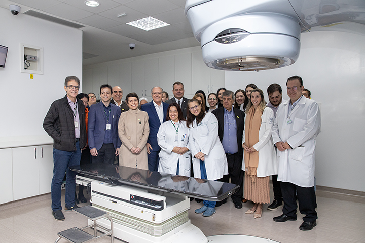 A imagem mostra um grupo de profissionais, incluindo médicos, técnicos e gestores, posando em uma sala de um hospital ou centro de saúde. Ao centro, há um equipamento de grande porte, identificado como um acelerador linear, usado para tratamentos de radioterapia. O ambiente é moderno, limpo e bem iluminado, reforçando a impressão de um espaço dedicado à tecnologia médica avançada.
