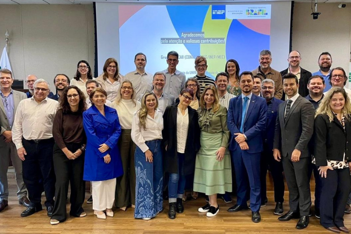 A imagem mostra um grupo de aproximadamente 30 pessoas posando para uma foto em um ambiente interno, provavelmente uma sala de conferência ou auditório. Ao fundo, há uma tela de projeção com uma apresentação exibida, indicando que o evento está relacionado ao INEP (Instituto Nacional de Estudos e Pesquisas Educacionais Anísio Teixeira), ao Ministério da Educação (MEC) e à avaliação da educação superior. O grupo é formado por homens e mulheres de diferentes idades, todos bem vestidos, em trajes formais e semiformal. Eles aparentam estar felizes, sorrindo para a foto, sugerindo que o evento foi positivo ou celebrativo. A maioria está de pé, enquanto alguns estão levemente agachados na frente para melhor se posicionar na imagem.