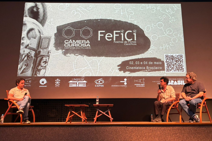 A imagem mostra um painel no FeFiCi – Festival de Filmes Científicos, realizado de 2 a 4 de maio na Cinemateca Brasileira. Três homens conversam no palco, com um banner projetado ao fundo destacando o tema do evento: “Câmera Curiosa – Ciência em Filmes”. O festival tem apoio de instituições como a Unifesp, Capes e o Governo Federal.
