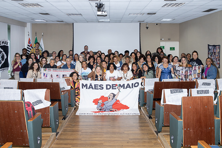 Imagem mostra participantes do evento reunidos à frente da mesa de abertura, segurando faixas e cartazes