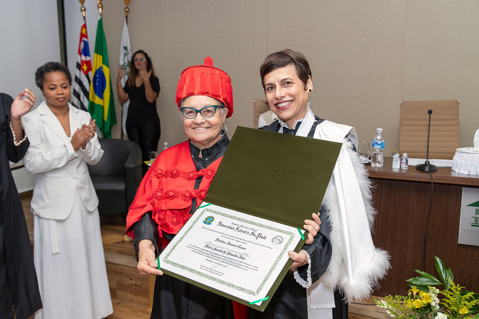 Na imagem, duas mulheres estão em destaque em uma cerimônia de entrega de título honorífico na Universidade Federal de São Paulo (Unifesp). À esquerda, uma senhora com cabelos grisalhos usa óculos e veste uma beca vermelha com detalhes bordados e um chapéu também vermelho. Ela segura um diploma verde e branco, sorrindo para a câmera. Ao lado dela, à direita, uma mulher com cabelo curto e castanho veste uma beca preta e branca, também sorrindo enquanto segura o outro lado do diploma. Ao fundo, há outras pessoas aplaudindo, e atrás delas, estão bandeiras do Brasil e do estado de São Paulo, além da bandeira da Unifesp.