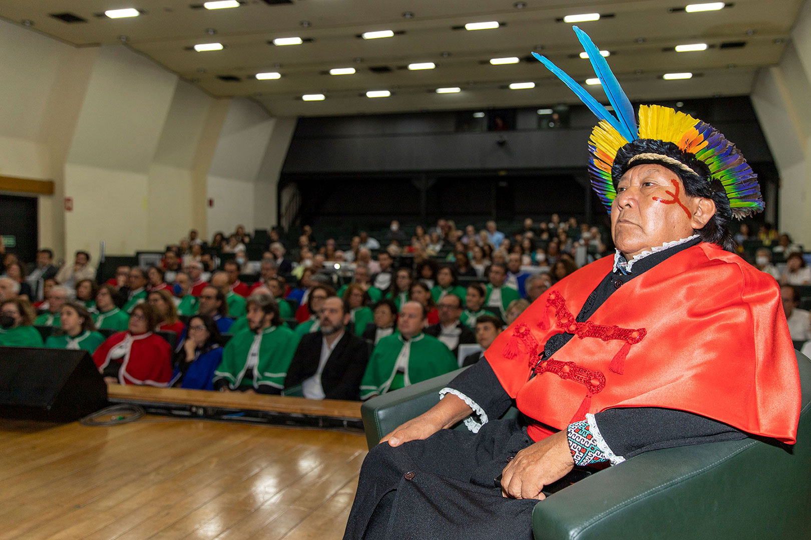 Na imagem, há um homem indígena em destaque, sentado em uma cadeira durante uma cerimônia de entrega de título honorífico na Universidade Federal de São Paulo (Unifesp). Ele veste uma beca vermelha por cima de suas roupas tradicionais e um cocar com penas coloridas de azul, amarelo, verde e preto. Seu rosto está pintado com tinta vermelha, em um padrão tradicional indígena. Ele tem uma expressão séria e está concentrado na cerimônia. Ao fundo, o auditório está cheio de pessoas sentadas, muitas delas vestindo becas verdes, vermelhas e azuis, assistindo atentamente à solenidade. A iluminação é suave e o ambiente é formal, com o foco principal sendo o homenageado indígena.