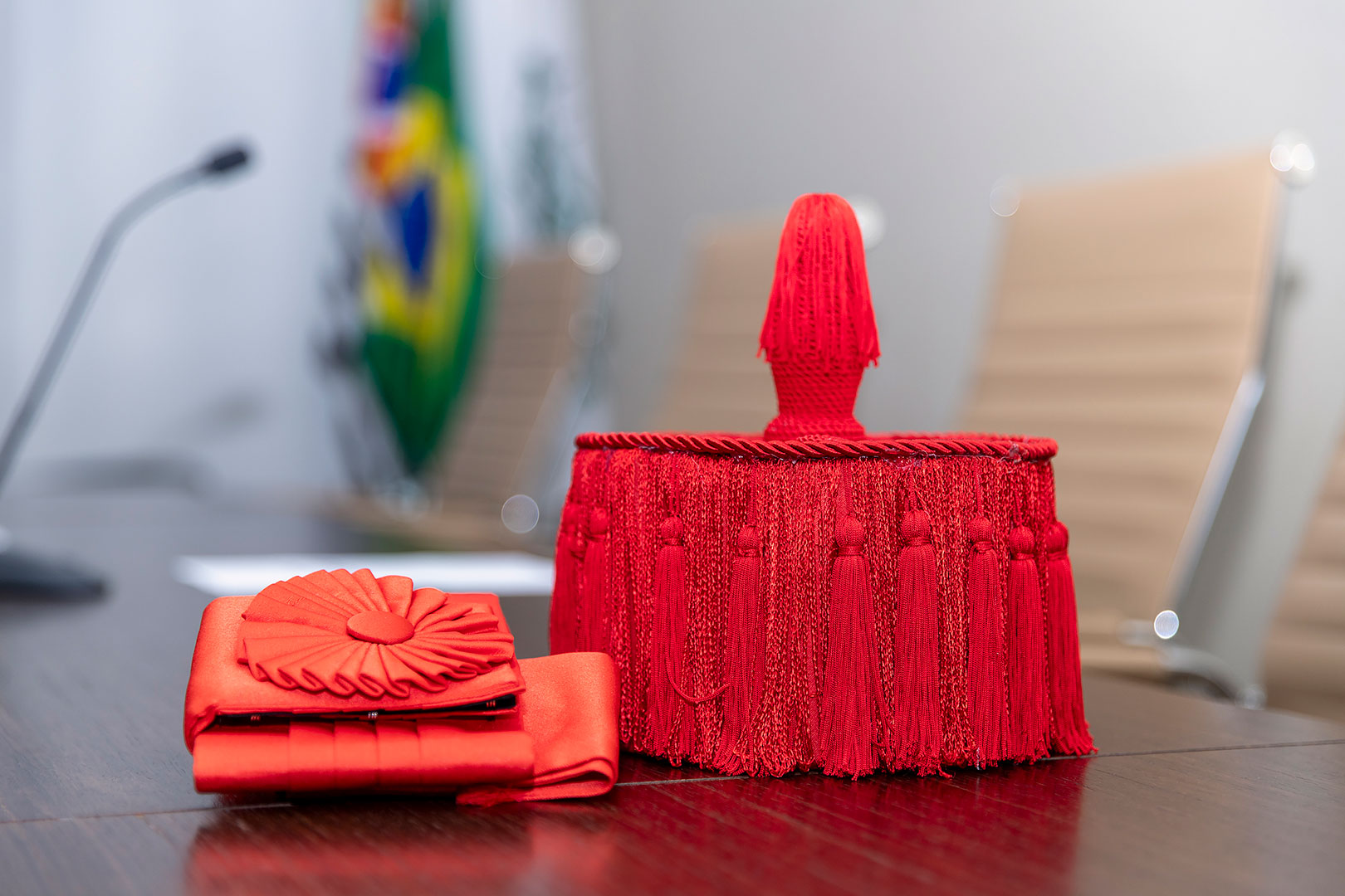 Imagem da faixa e chapéu que são usados no cerimônia de titulação - eles são vermelhos e estão sob uma mesa, ao fundo está a bandeira do Brasil